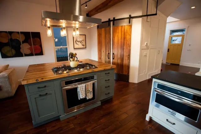 A modern rustic kitchen featuring a sage green island with a wood countertop, stainless steel appliances, and sliding barn doors.